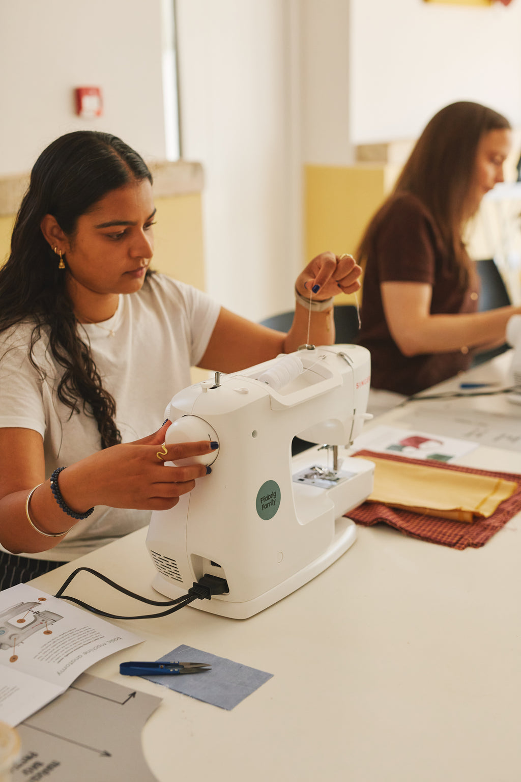 A woman in a white t-shirt operates a white sewing machine at a table in a bright indoor workshop, with another person blurred in the background.
