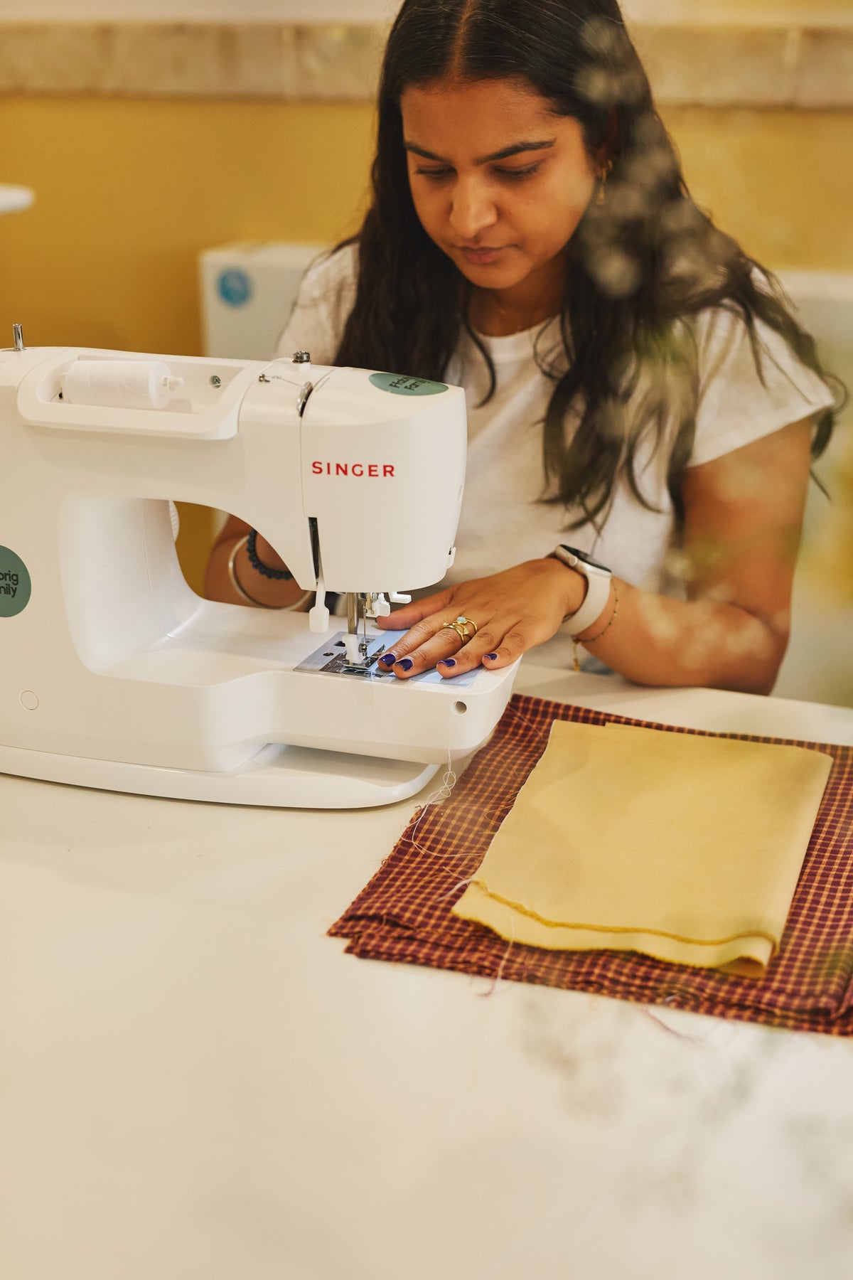 A woman operating a white Singer sewing machine at a table, with fabric pieces on the table in front of her.