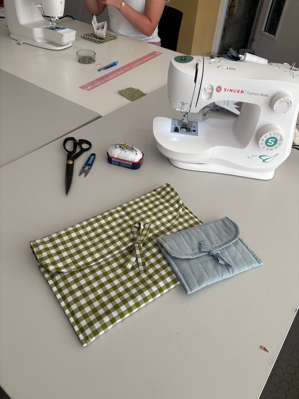 Two handmade quilted fabric pouches with bow details, one in olive green and white gingham check and the other in light blue, displayed on a sewing table alongside a white Singer sewing machine, scissors, and a pin cushion.