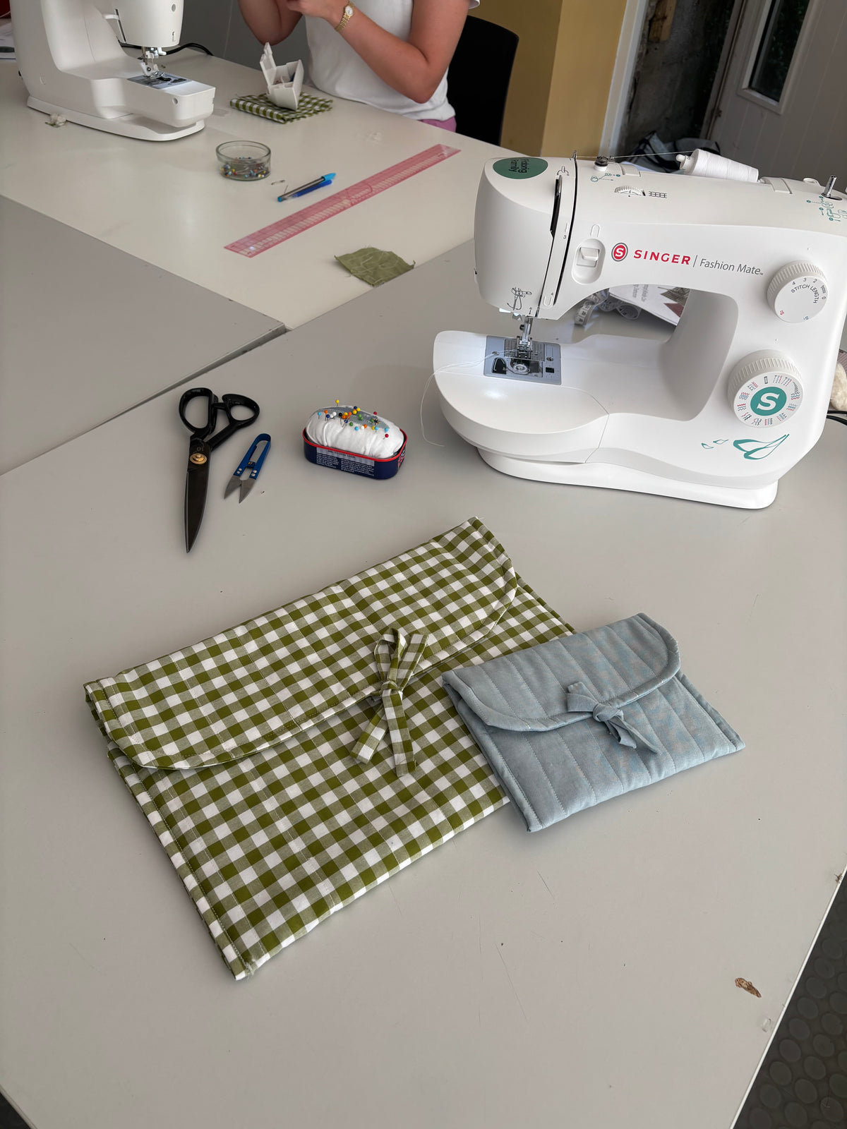 Two handmade quilted fabric pouches with bow details, one in olive green and white gingham check and the other in light blue, displayed on a sewing table alongside a white Singer sewing machine, scissors, and a pin cushion.