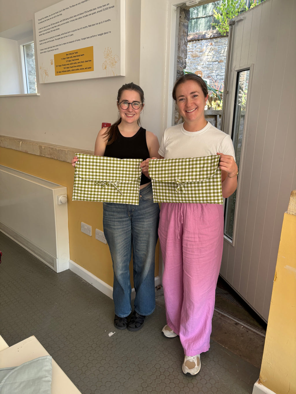 Two women standing indoors holding matching green and white gingham check pocket pouches with embroidered bow details. The woman on the left wears a black tank top and blue jeans, and the woman on the right wears a white t-shirt and pink pants.