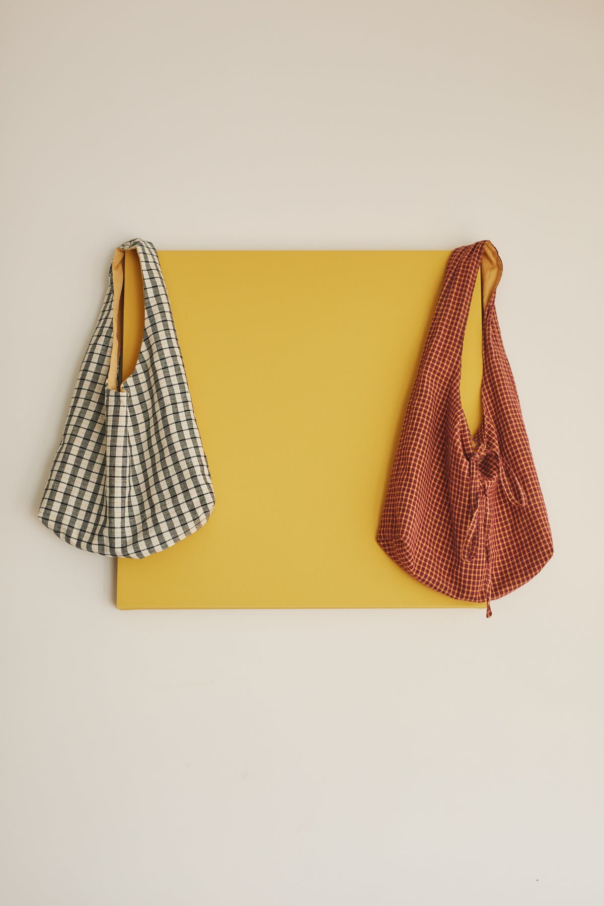 Two fabric shoulder bags hanging from a yellow rectangular board against a plain off-white wall. The left bag is black and white gingham with a mustard yellow strap, and the right bag is a rust-red and white gingham with a yellow strap.