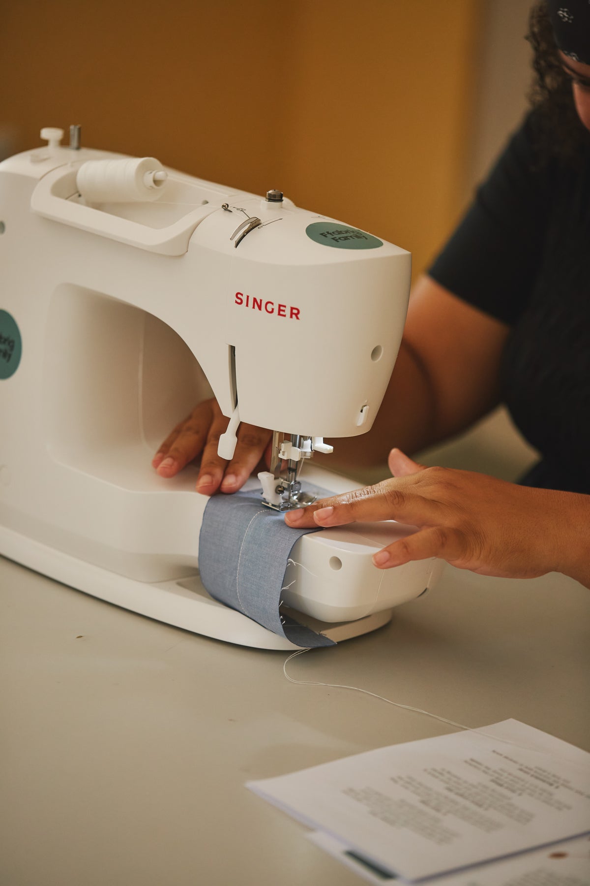 A white Singer sewing machine being operated by a person whose hands and partial torso are visible, with blue-gray fabric under the needle on a tabletop.