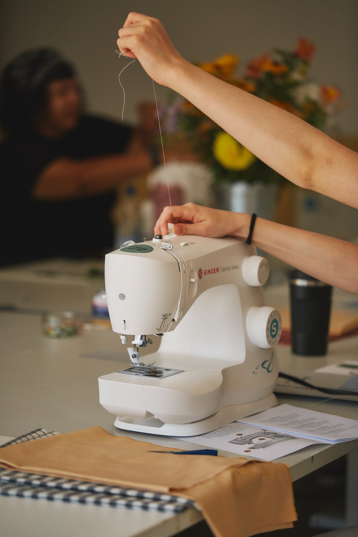 A white Singer sewing machine on a table in a workshop setting, with a person's hands adjusting the machine and another person blurred in the background.