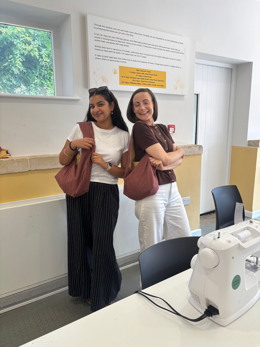 Two women standing in a bright room, each holding a dusty rose textured tote bag. They are positioned in front of a white wall with a window, informational sign, and a sewing machine on a table in the foreground.