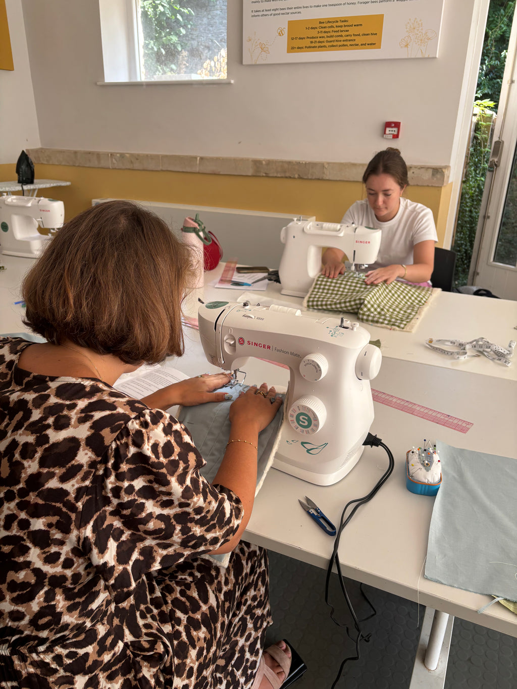 Two women sewing at white sewing machines in a bright room with a yellow accent wall. The woman in the foreground wears a leopard print top and is seen from behind, while the woman in the background wears a white t-shirt.