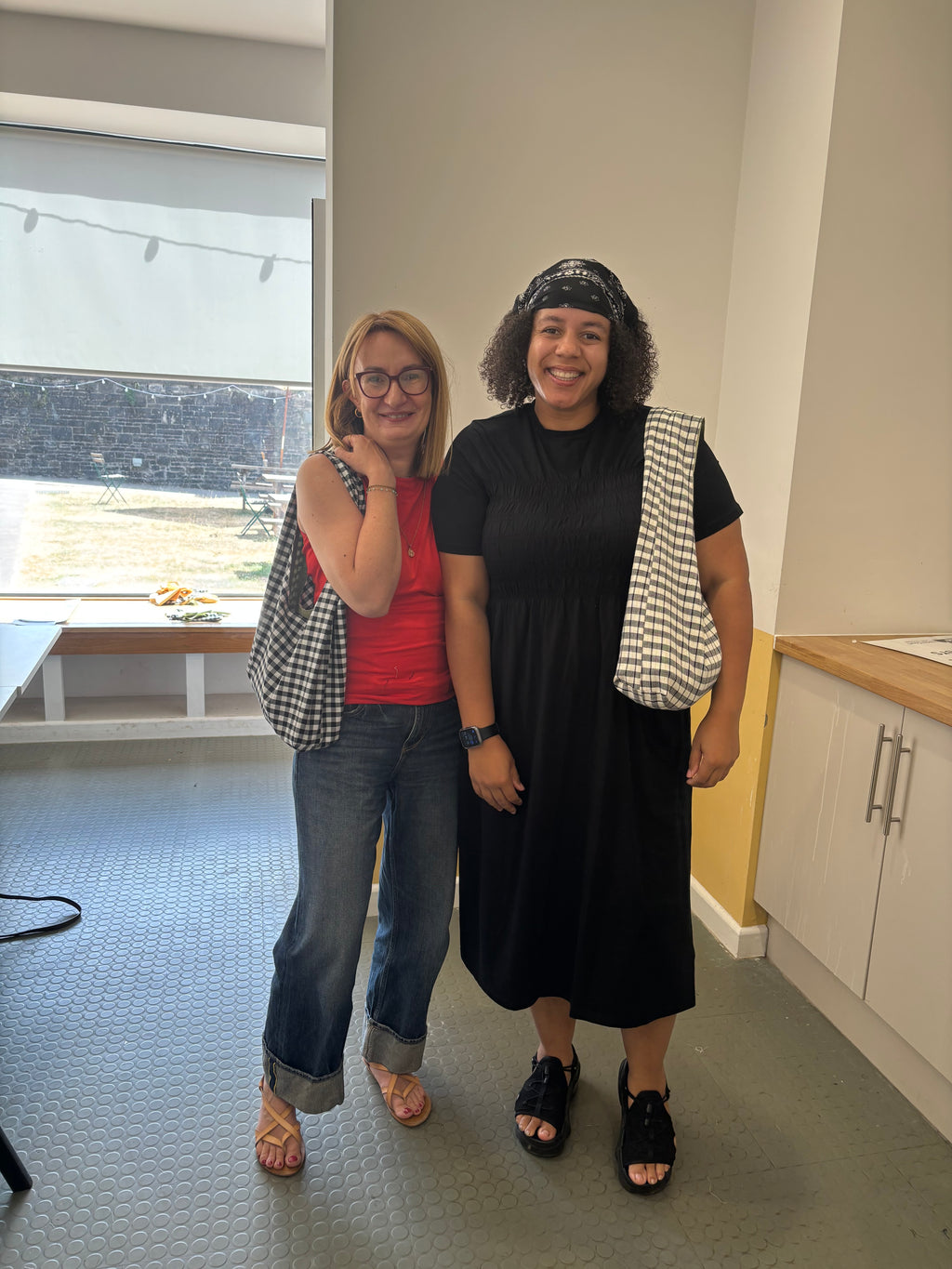 Two women standing together in a bright indoor space, each carrying a black and white checkered tote bag. The woman on the left wears a red top and blue jeans, while the woman on the right wears a black dress and black sandals.