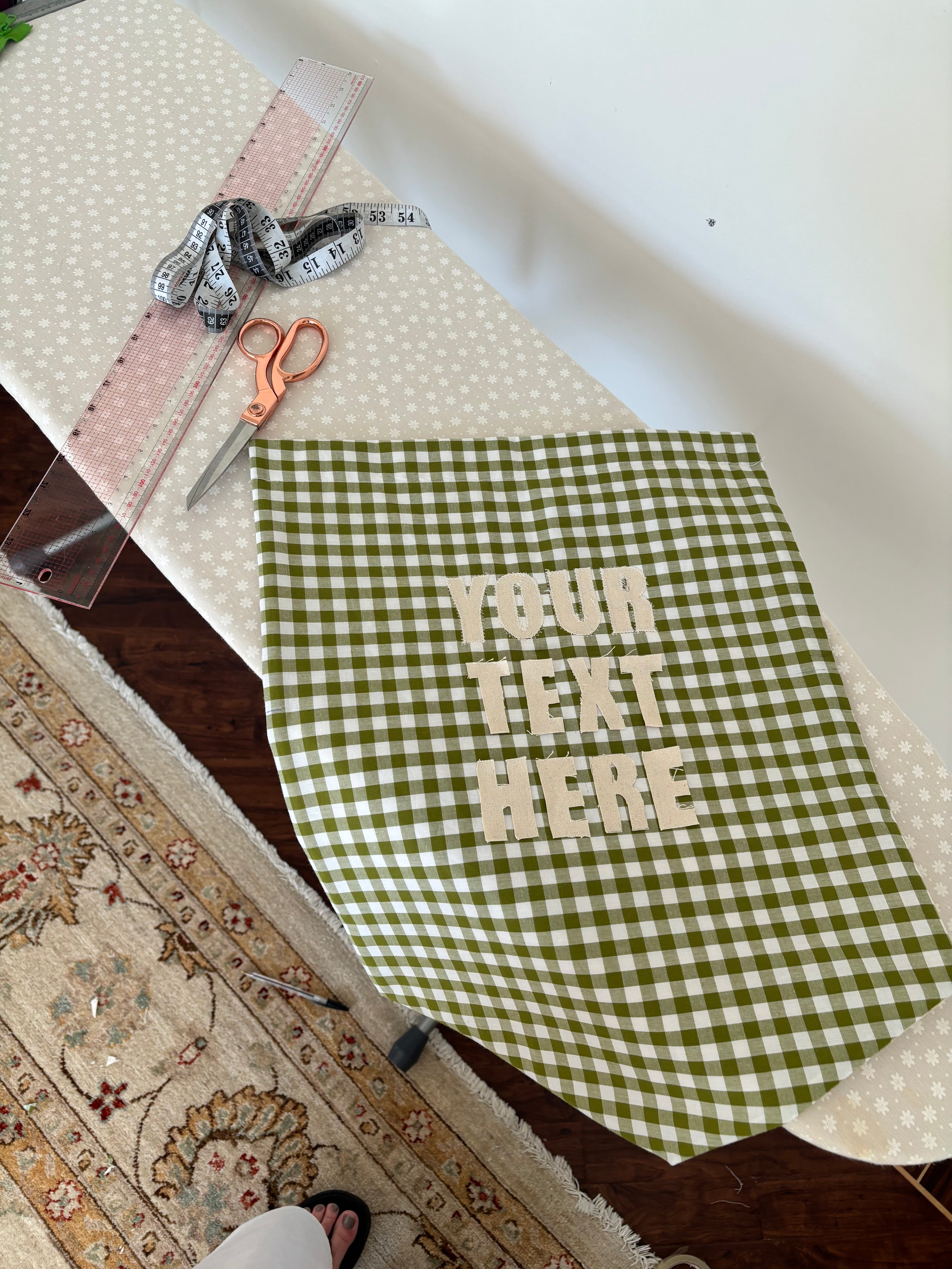 A green and white gingham fabric apron or bib with 'YOUR TEXT HERE' printed in beige, laid on a white polka-dot ironing board with measuring tools and scissors nearby.